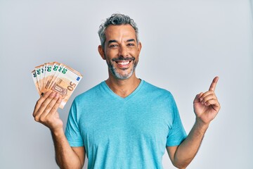 Middle age grey-haired man holding 50 euro banknotes smiling happy pointing with hand and finger to...