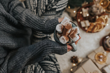 Young woman in warm winter home pants sits on a soft blanket. lady enjoying hot cocoa with marshmallow and cinnamon.Still life details in home interior. 