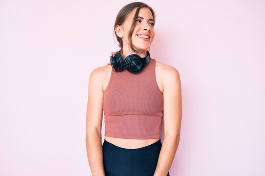 Beautiful young caucasian woman wearing gym clothes and using headphones looking away to side with smile on face, natural expression. laughing confident.