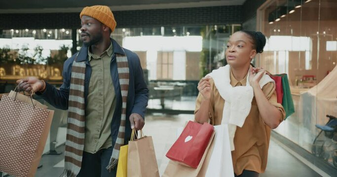 Excited joyful black young couple of stylish people running in hallway of shopping mall with bags enjoying holiday sale shopping on black friday.