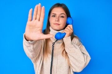 Beautiful caucasian woman speaking on vintage telephone with open hand doing stop sign with serious and confident expression, defense gesture