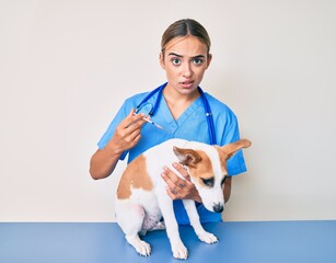 Young beautiful blonde veterinarian woman putting vaccine to puppy dog skeptic and nervous,...