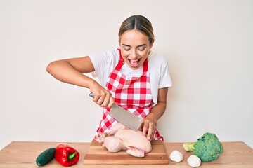 Fototapeta premium Young beautiful blonde woman wearing cook apron cutting chicken smiling and laughing hard out loud because funny crazy joke.