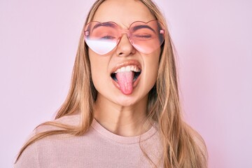 Young beautiful blonde woman wearing heart shaped sunglasses sticking tongue out happy with funny...