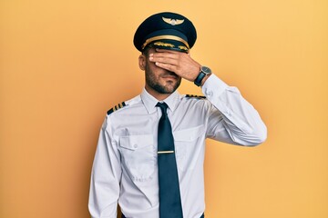 Handsome hispanic man wearing airplane pilot uniform covering eyes with hand, looking serious and sad. sightless, hiding and rejection concept