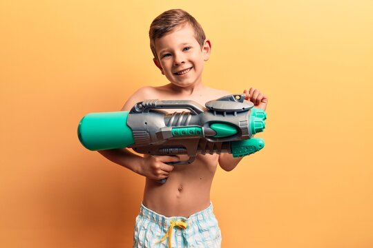 Cute blond kid wearing swimwear holding water gun screaming proud, celebrating victory and success very excited with raised arm