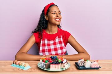 Young african american girl wearing baker uniform sitting on the table with sweets looking to side, relax profile pose with natural face and confident smile.