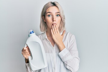 Young blonde girl holding detergent bottle covering mouth with hand, shocked and afraid for...