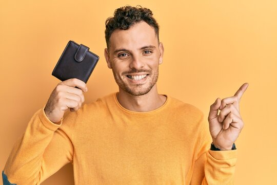 Young hispanic man holding leather wallet smiling happy pointing with hand and finger to the side