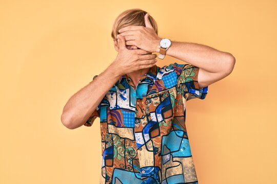 Young blond man wearing summer shirt covering eyes and mouth with hands, surprised and shocked. hiding emotion