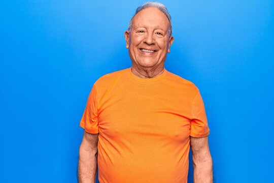 Senior handsome grey-haired man wearing casual t-shirt standing over blue background with a happy and cool smile on face. Lucky person.