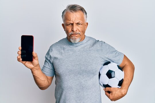 Middle age grey-haired man holding football ball showing smartphone skeptic and nervous, frowning upset because of problem. negative person.