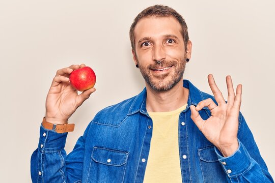 Young handsome man holding red apple doing ok sign with fingers, smiling friendly gesturing excellent symbol