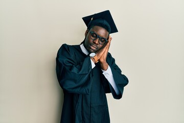 Handsome black man wearing graduation cap and ceremony robe sleeping tired dreaming and posing with hands together while smiling with closed eyes.