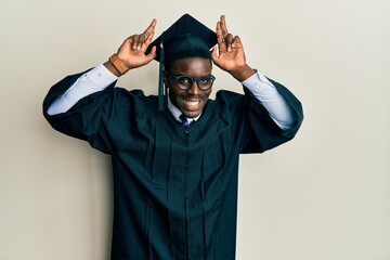 Handsome black man wearing graduation cap and ceremony robe posing funny and crazy with fingers on...