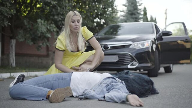 Stressed young blond woman checking pulse of body hit by car outdoors. Portrait of frustrated young Caucasian female driver and dead or injured victim on summer road. Over speeding concept.