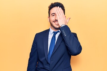 Young hispanic man wearing suit covering one eye with hand, confident smile on face and surprise emotion.