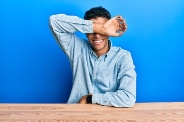 Young handsome african american man wearing casual clothes sitting on the table covering eyes with arm smiling cheerful and funny. blind concept.