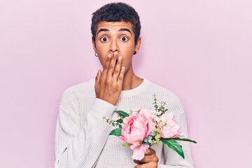 Young african amercian man holding flowers covering mouth with hand, shocked and afraid for...