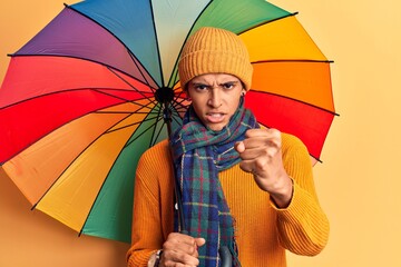 Young african amercian man holding colorful umbrella annoyed and frustrated shouting with anger,...