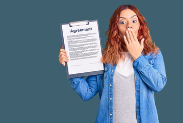 Young latin woman holding clipboard with agreement document covering mouth with hand, shocked and...