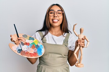 Young latin woman artist holding palette and manikin sticking tongue out happy with funny expression.