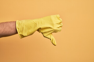 Hand of caucasian young man with cleaning glove over isolated yellow background doing thumbs down...