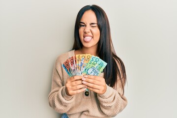 Young beautiful hispanic girl holding australian dollars sticking tongue out happy with funny...