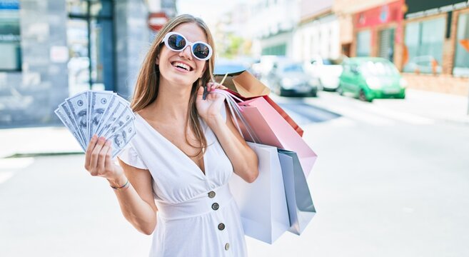 Young blonde woman smiling happy holding shopping bags and dollars banknotes at street of city