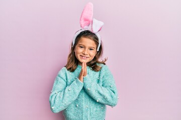Little beautiful girl wearing cute easter bunny ears praying with hands together asking for...