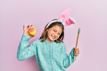 Little beautiful girl wearing cute easter bunny ears holding colored egg winking looking at the...