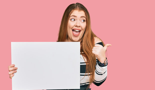 Young beautiful redhead woman holding blank empty banner pointing thumb up to the side smiling happy with open mouth
