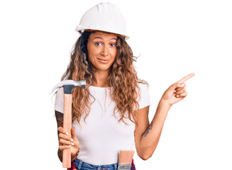Young hispanic woman with tattoo wearing hardhat and builder clothes holding hammer smiling happy pointing with hand and finger to the side
