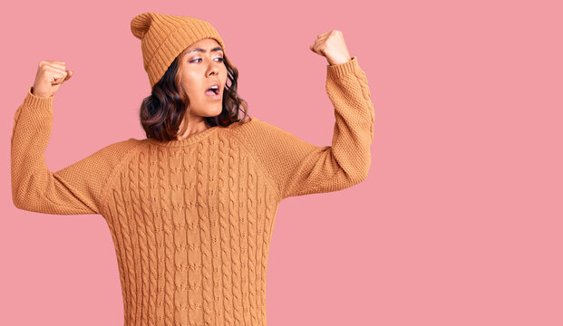 Young beautiful mixed race woman wearing wool sweater and winter hat showing arms muscles smiling proud. fitness concept.
