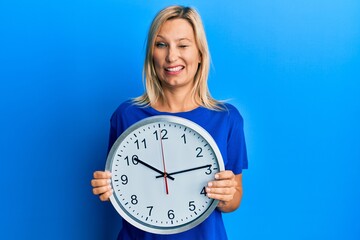 Beautiful middle age blonde woman holding big clock winking looking at the camera with sexy...