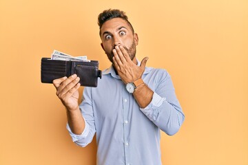 Handsome man with beard holding wallet with dollars covering mouth with hand, shocked and afraid...