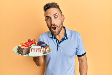 Handsome man with beard holding cake slices scared and amazed with open mouth for surprise, disbelief face