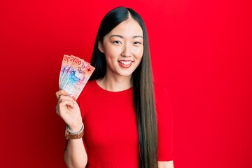 Young chinese woman holding 20 swiss franc banknotes looking positive and happy standing and smiling with a confident smile showing teeth