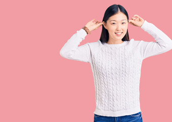 Young beautiful chinese woman wearing casual sweater smiling pulling ears with fingers, funny...