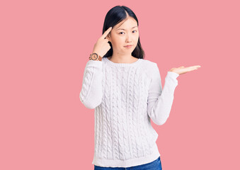 Young beautiful chinese woman wearing casual sweater confused and annoyed with open palm showing copy space and pointing finger to forehead. think about it.