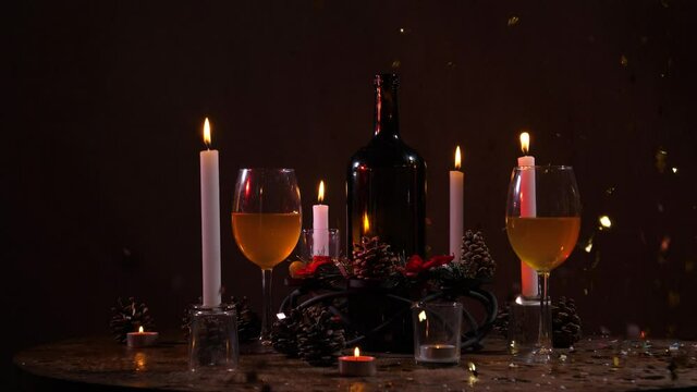 On a dark, brown background, a festive New Year's still life is depicted in the form of a bottle of wine, burning candles, filled glasses and fir cones in front of New Year's confetti pouring down.