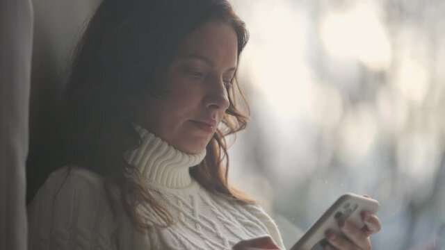Attractive pregnant woman holding smart phone sitting by window at home, pregnancy and motherhood.