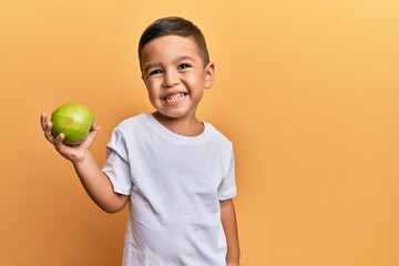 Adorable latin toddler smiling happy holding green apple looking to the camera over isolated yellow background.