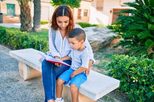 Adorable latin mother and son sitting on the bench and reading book at the park.