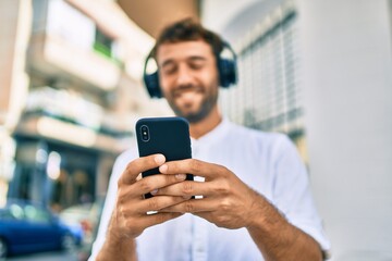 Handsome man with beard wearing casual white shirt on a sunny day smiling happy outdoors using smartphone listening to music wearing headphones