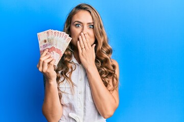 Young blonde girl holding 10 colombian pesos banknotes covering mouth with hand, shocked and afraid for mistake. surprised expression