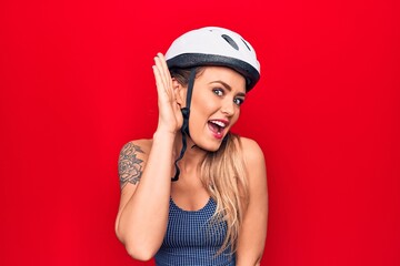 Young beautiful blonde cyclist woman wearing bike security helmet over red background smiling with hand over ear listening and hearing to rumor or gossip. Deafness concept.