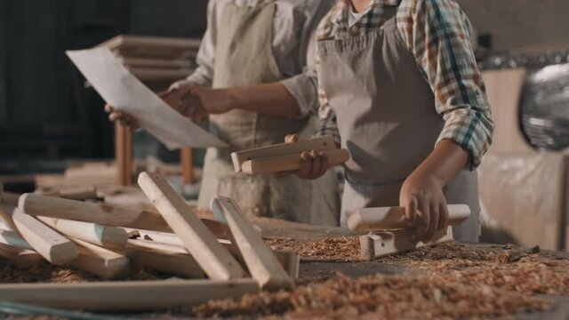 Midsection footage of unrecognizable man with little boy in beige aprons making diy project gathering wooden slats together at carpentry table