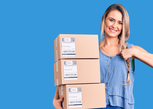 Young beautiful blonde woman holding delivery boxes pointing finger to one self smiling happy and proud