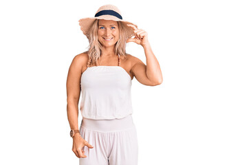 Obraz na płótnie Canvas Middle age fit blonde woman wearing summer hat smiling and confident gesturing with hand doing small size sign with fingers looking and the camera. measure concept.